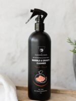 Cleaning Mama plant-based Fresh Scent Marble and Granite Cleaner in a black spray bottle displayed on a wooden board next to a plant and a white microfiber cloth.