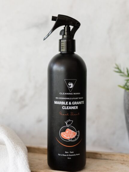 Cleaning Mama plant-based Fresh Scent Marble and Granite Cleaner in a black spray bottle displayed on a wooden board next to a plant and a white microfiber cloth.
