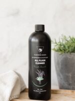 Cleaning Mama non-toxic, plant-based Fresh Scent All Floor Cleaner in a black spray bottle displayed on a wooden board next to a plant and a white microfiber cloth.