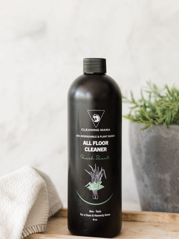 Cleaning Mama non-toxic, plant-based Fresh Scent All Floor Cleaner in a black spray bottle displayed on a wooden board next to a plant and a white microfiber cloth.