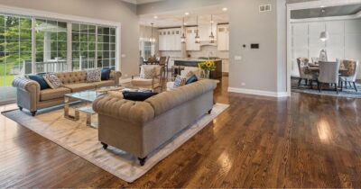 Bright open living room with polished hardwood floors, two sofas on a rug, a glass coffee table, and a dining area and kitchen in the background.